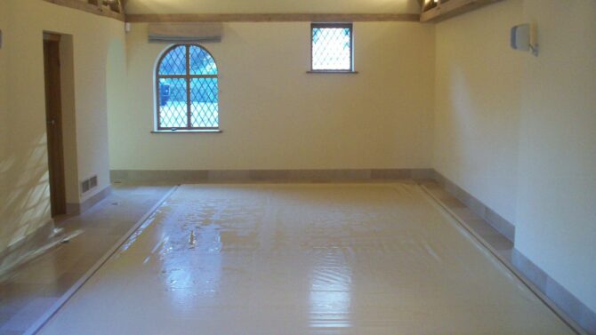 Stone AQM Rectangular indoor pool covered by a taut beige protective sheet, recessed into tiled floor; room features cream walls, exposed wooden roof beams, two diamond‑pane windows and soft overhead lighting.