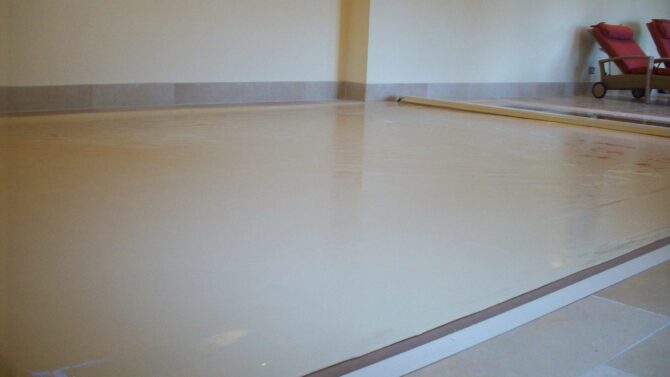 Stone AWM A large rectangular indoor pool covered with taut beige vinyl, recessed into a tiled floor; two red padded lounge chairs and a small leaded window sit against cream-colored walls.