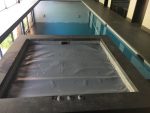 A square spa hot tub covered by a grey tarp sits empty in the foreground while a long indoor swimming pool stretches behind, surrounded by tiled deck and tall windows.
