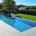 A rectangular infinity pool reflects a tree and blue sky, bordered by pale tiled terrace with lounge chairs and umbrellas, adjacent to a manicured green lawn and hedge-backed suburban houses.