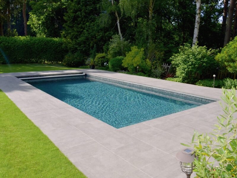 Rectangular in-ground swimming pool ripples with clear blue water, bordered by wide gray tiles and neat green lawn, flanked by potted plants, shrubs and tall trees in a backyard.
