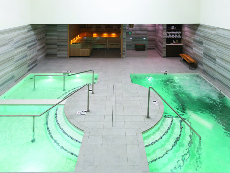 Two adjacent spa pools glowing green, bubbling and gently circulating, flanking a tiled central walkway with metal handrails; background shows glass-doored sauna and towel shelf. Text visible: 1.6, 4.
