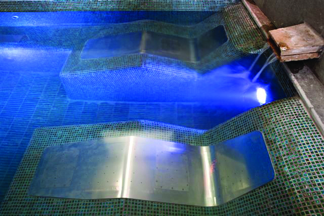 Wide Massage Bed – 400mm wide – Concrete Two stainless-steel lounge panels lie submerged as water pours from a small spout and blue LED lights illuminate the mosaic-tiled pool/spa edge.
